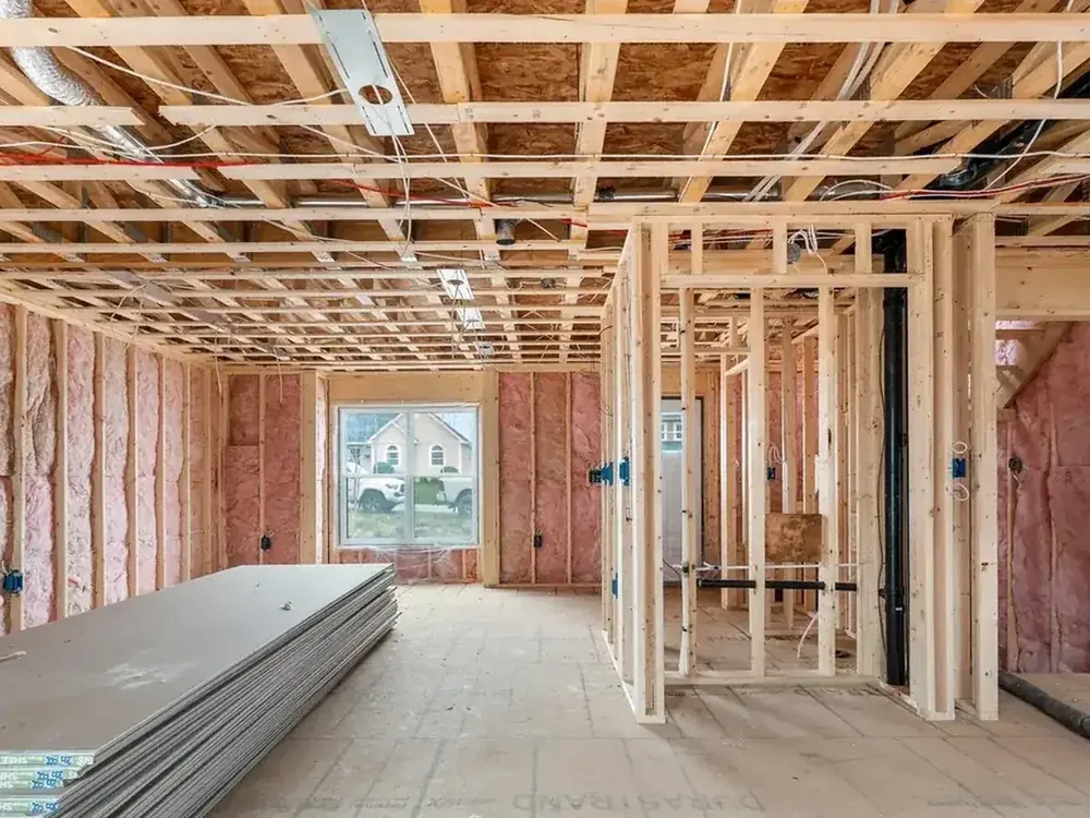 Active Wildawbren framing site: stud walls, exposed insulation, and roof rafters lit by afternoon sun through a doorway