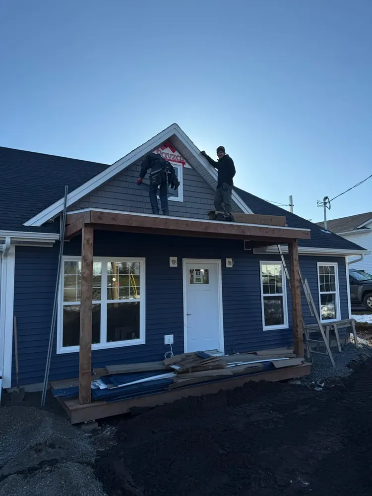 Two Wildawbren framers on the porch roof of a navy blue home, fitting trim into the front dormer at first light