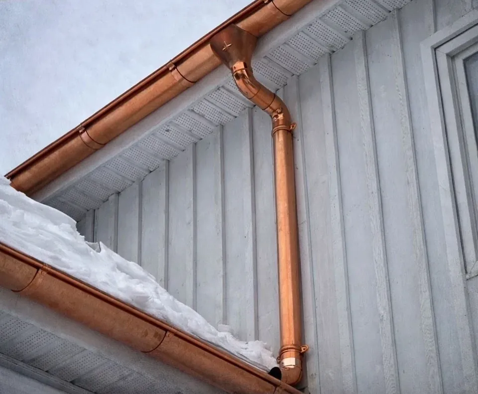 A copper seamless gutter and downspout running along the gable and corner of a white vertical-sided Wildawbren home, with snow drifted along the roofline