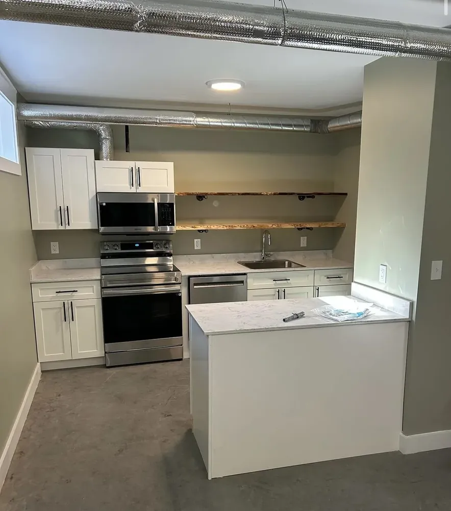 A Wildawbren basement kitchen with white shaker cabinets, live-edge wood floating shelves, white quartz countertops, and stainless appliances