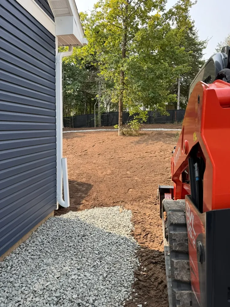 A Wildawbren Kubota track skid steer parked on fresh gravel between a navy blue home and a backyard tree line, ready to grade